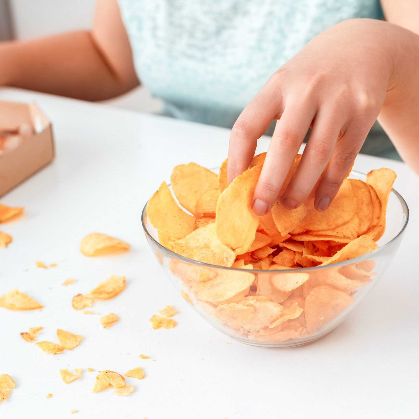 Young chubby woman sitting at table in kitchen with box of pizza breaking diet eating potato chip from bowl close-up